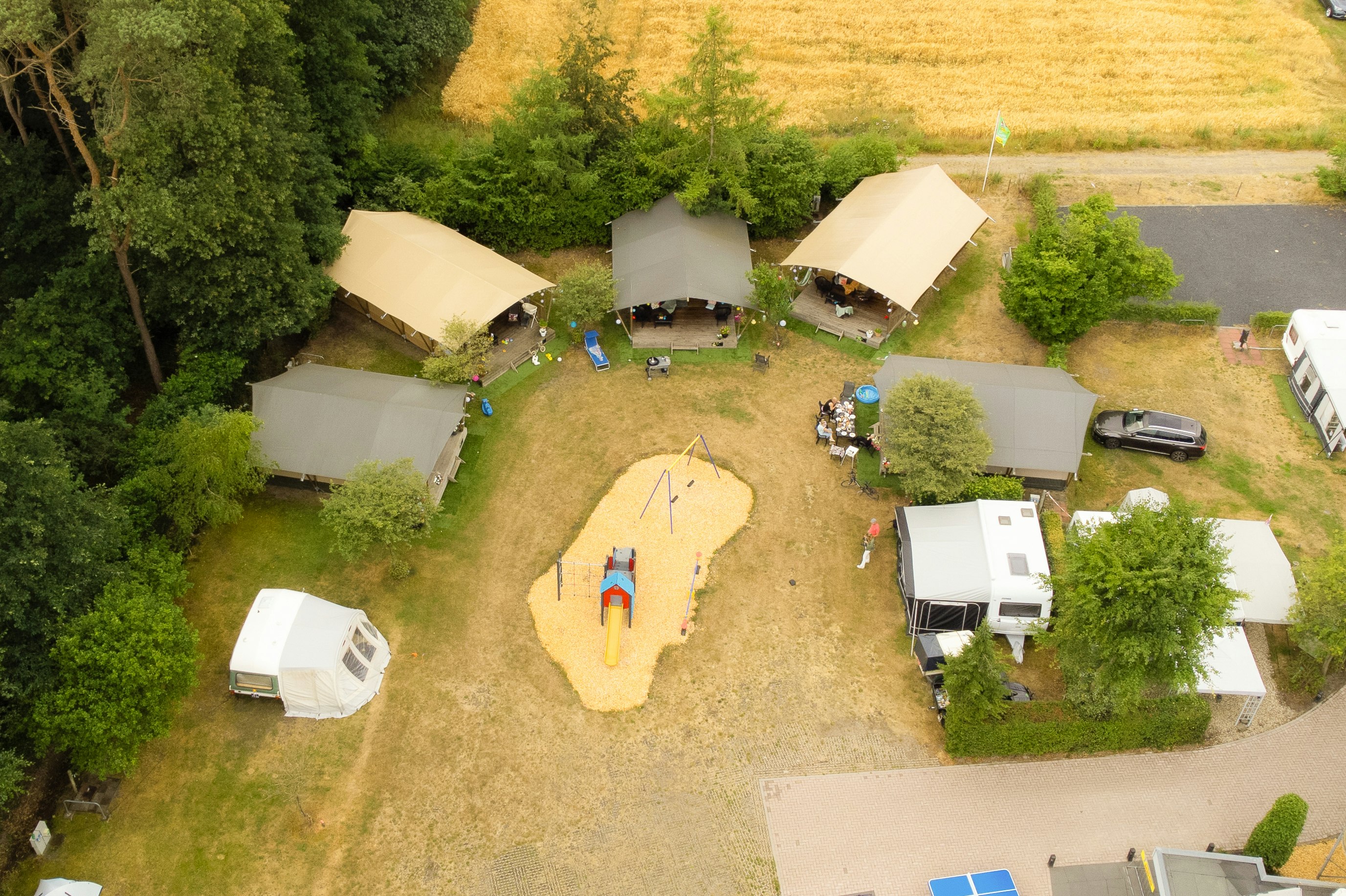 Luftaufnahme eines Campingplatzes mit mehreren Zelten, Wohnwagen und einem kleinen Spielbereich auf dem Rasen.