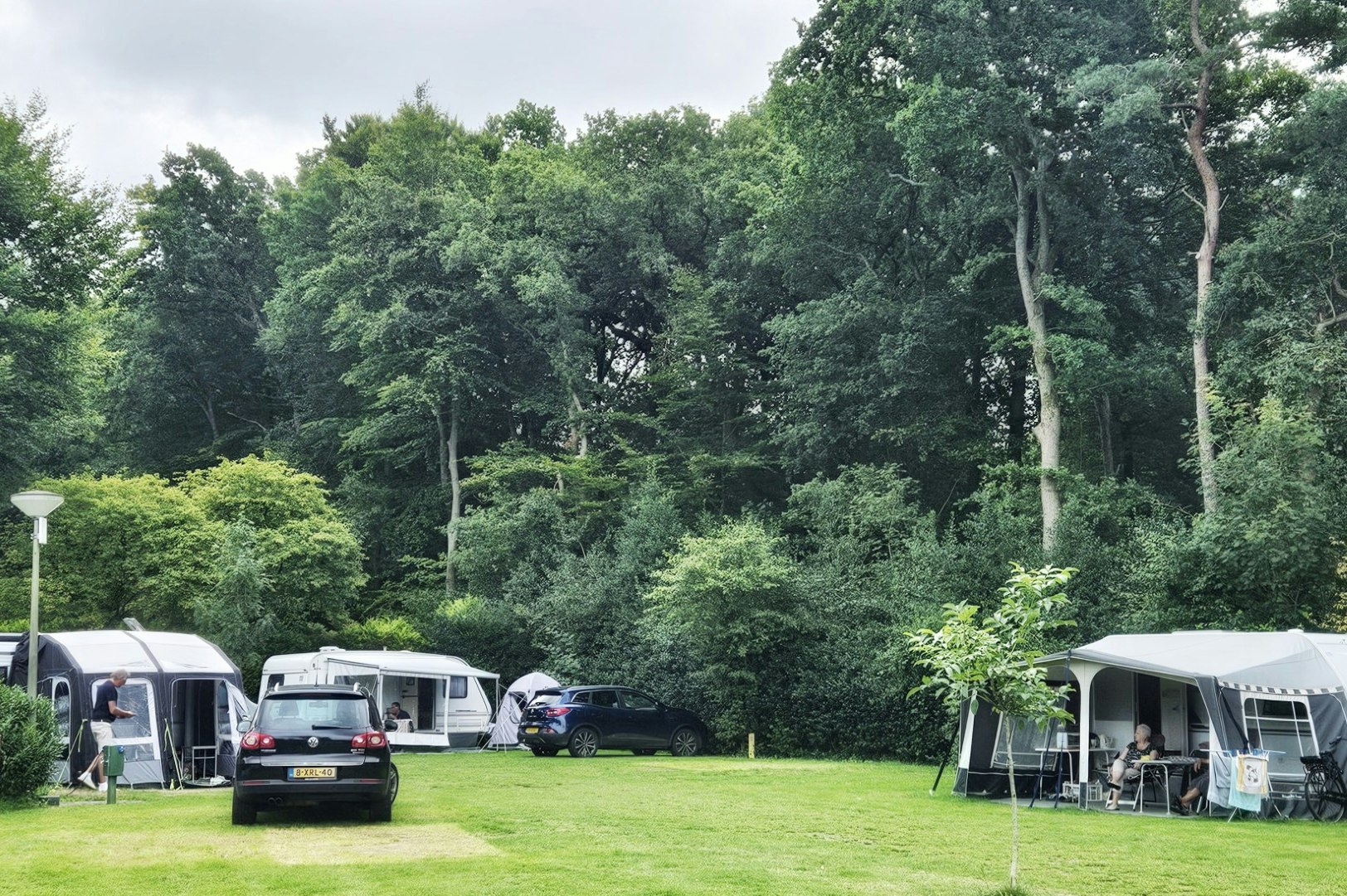 Campingplatz mit Wohnwagen und Zelten auf einer Rasenfläche, Bäume im Hintergrund und ein Auto auf dem Stellplatz.