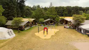 Campingplatz mit mehreren Zelten, kleinen Holzhütten und einem Spielplatz mit Rutsche auf einer Wiese.