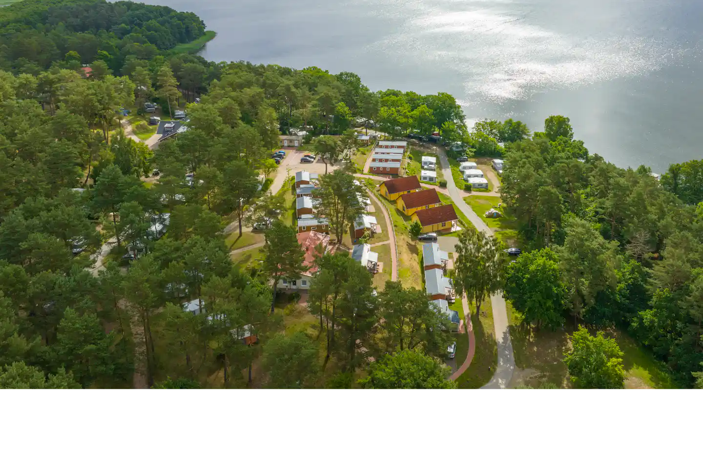 Luftaufnahme eines Campingplatzes zwischen Wäldern, mit Wohnwagen und Wegen entlang eines großen Sees.