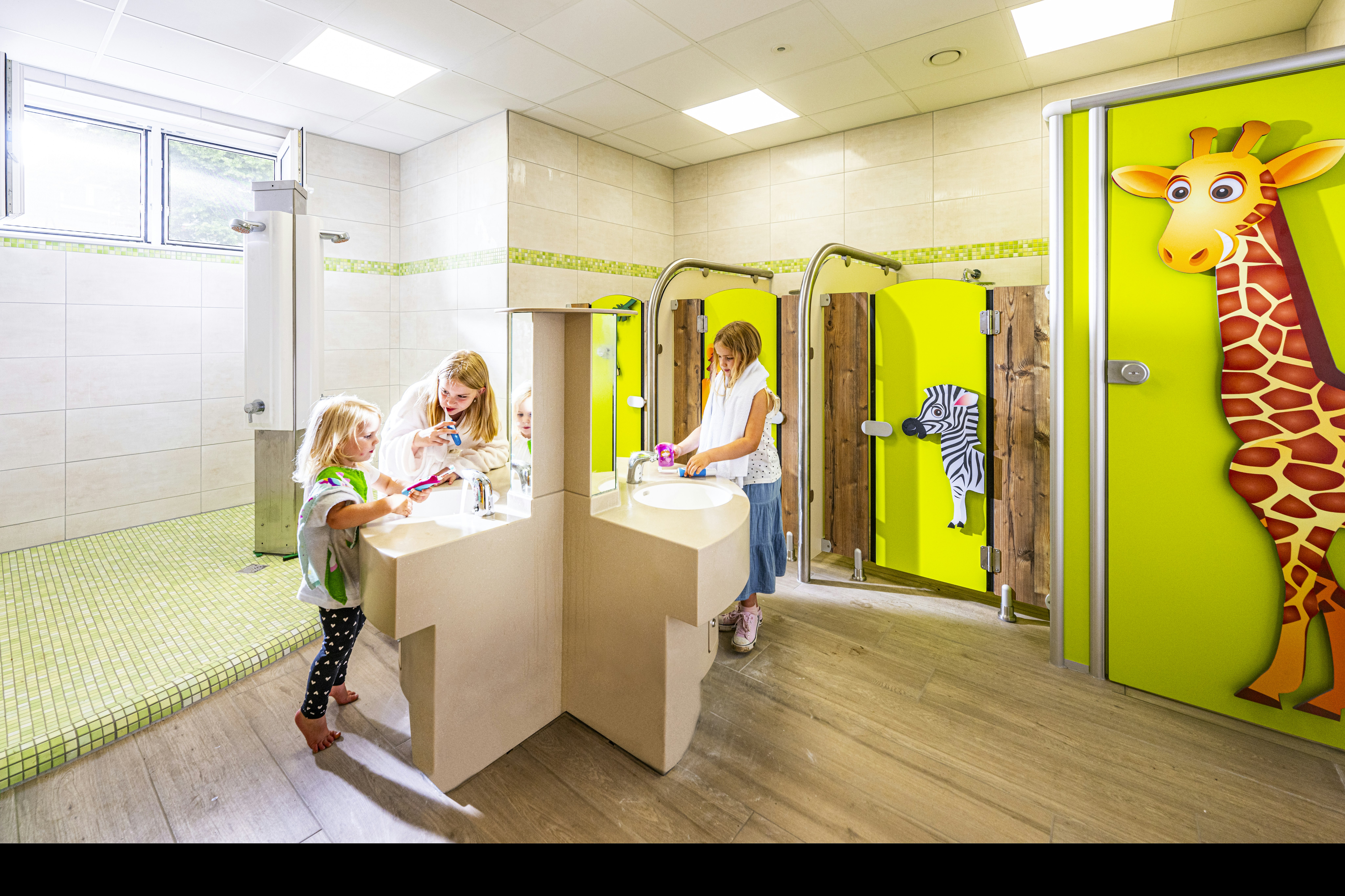 Kinderwaschbecken im hellen Sanitärraum, grüne Türen und ein großes Giraffenbild an der Wand.