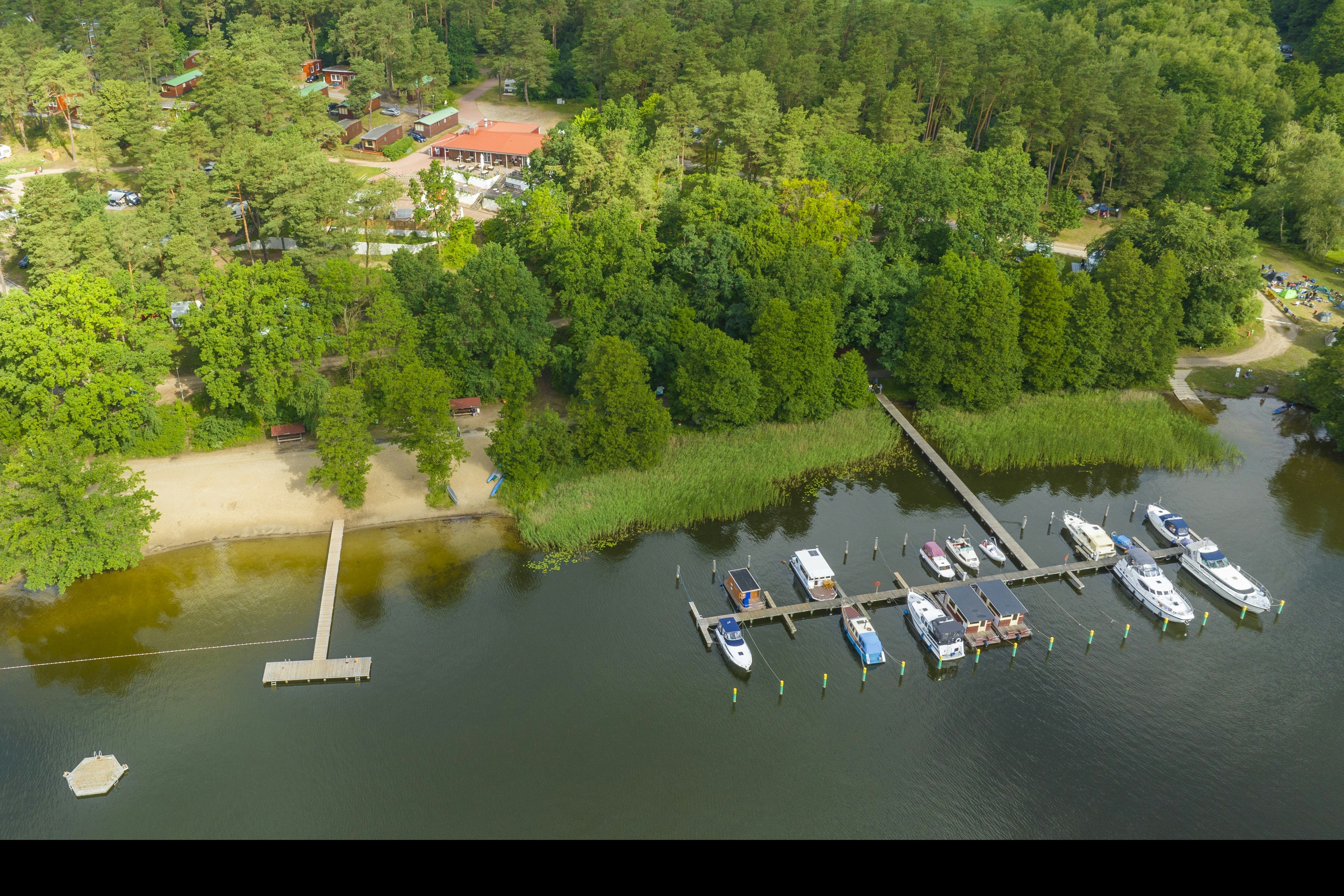 Luftaufnahme eines Campingplatzes am Fluss mit Steg, Booten und Stellplätzen im Grünen, umgeben von Bäumen und Wegen.