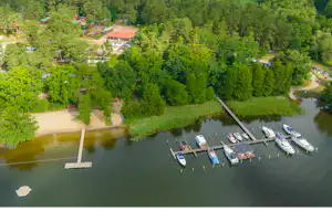 Luftaufnahme eines Campingplatzes am Fluss mit Steg, Booten und Stellplätzen im Grünen, umgeben von Bäumen und Wegen.