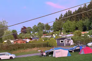 Campingplatz mit mehreren Zelten, Wohnwagen und kleinen Häuschen im Grünen, im Hintergrund Bäume und ein Zaun.