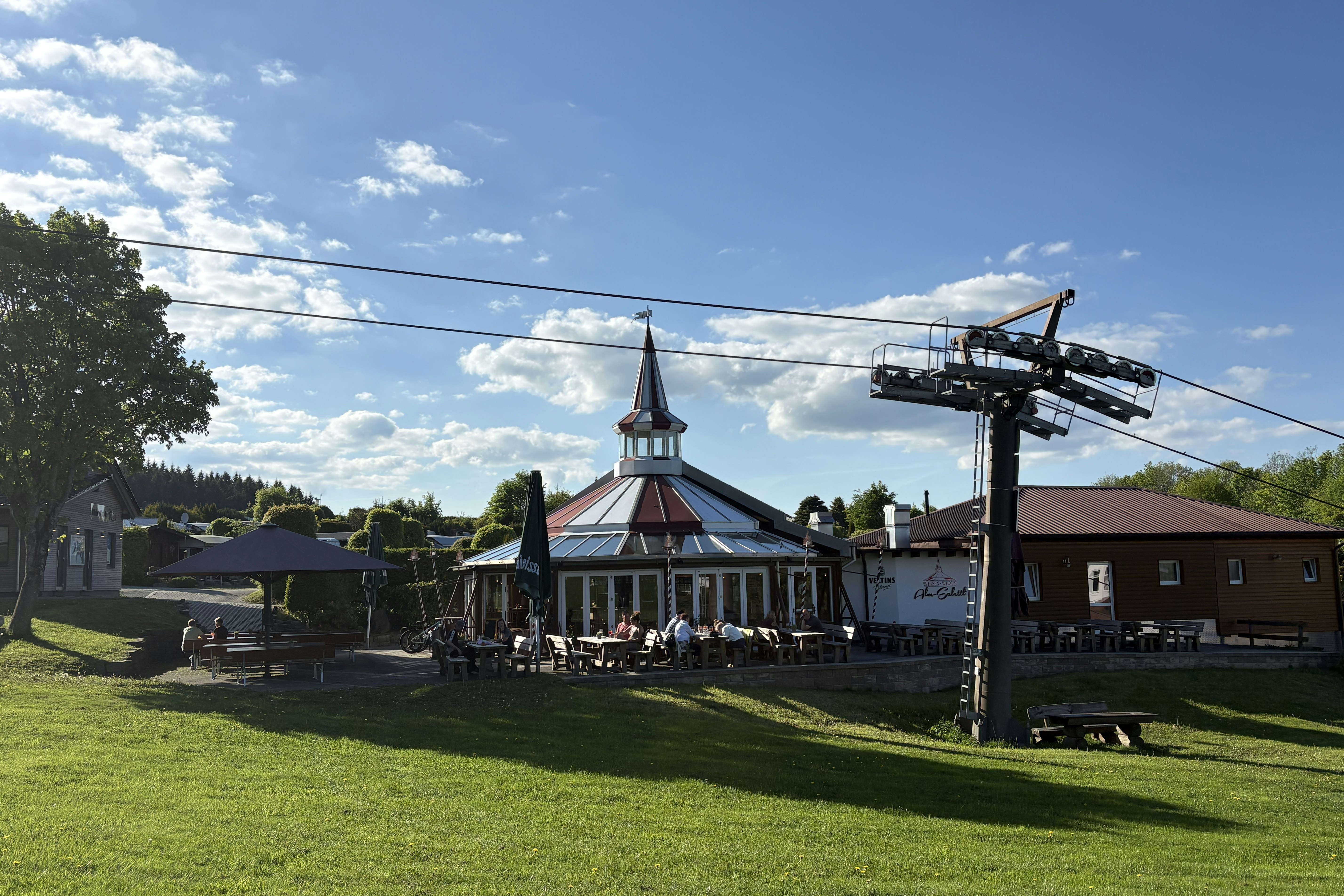 Campingplatz mit Rasenfläche, Pavillon mit Glockenturm und Sitzbereich, daneben ein großer Kran und Wohnwagen am Rand.