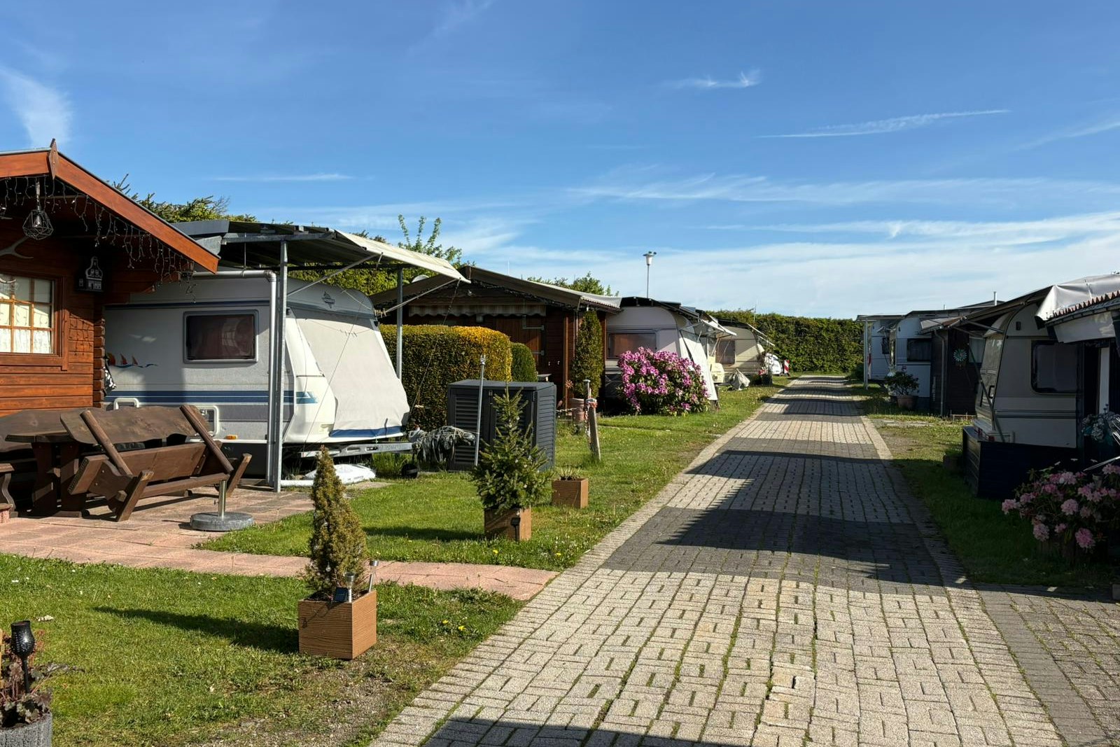 Reihen von Wohnwagen und kleinen Gartenparzellen mit gepflastertem Weg, Rasen und Hecken vor einem Wohnwagen.