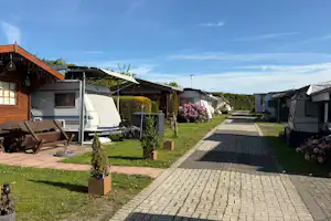 Reihen von Wohnwagen und kleinen Gartenparzellen mit gepflastertem Weg, Rasen und Hecken vor einem Wohnwagen.