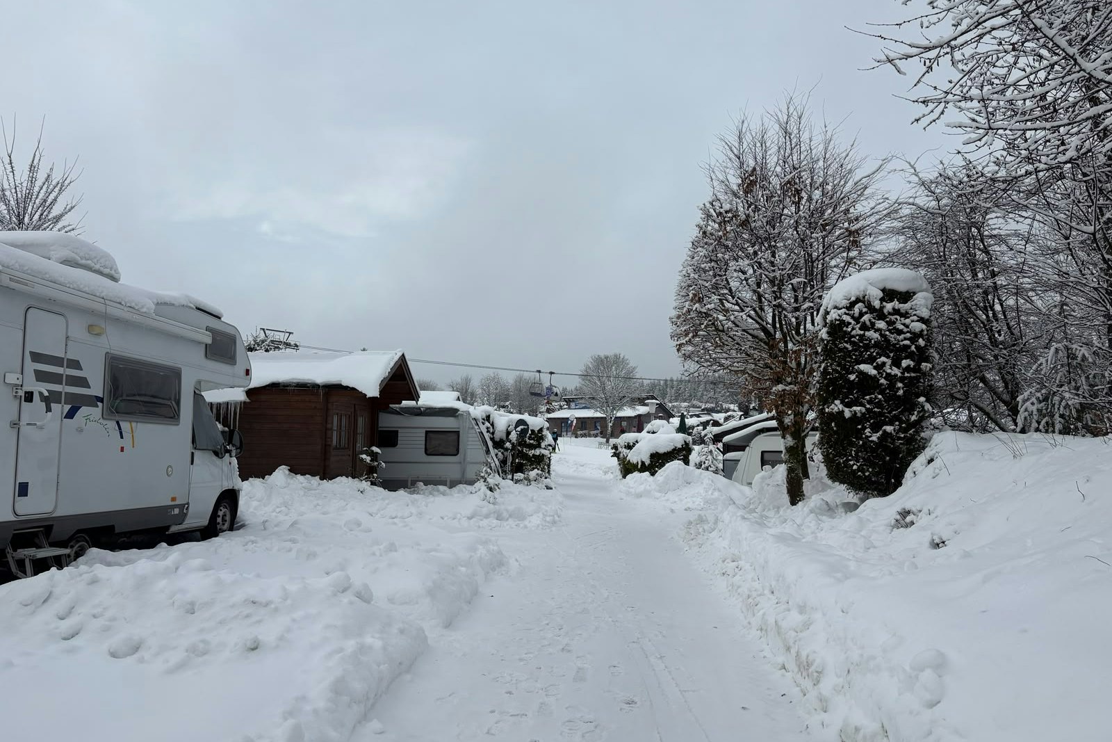 Wohnwagen und Wohnmobil auf einem verschneiten Campingplatz, mit Hecken, Bäumen und einem schmalen Weg zwischen den Stellplätzen.