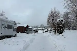 Wohnwagen und Wohnmobil auf einem verschneiten Campingplatz, mit Hecken, Bäumen und einem schmalen Weg zwischen den Stellplätzen.