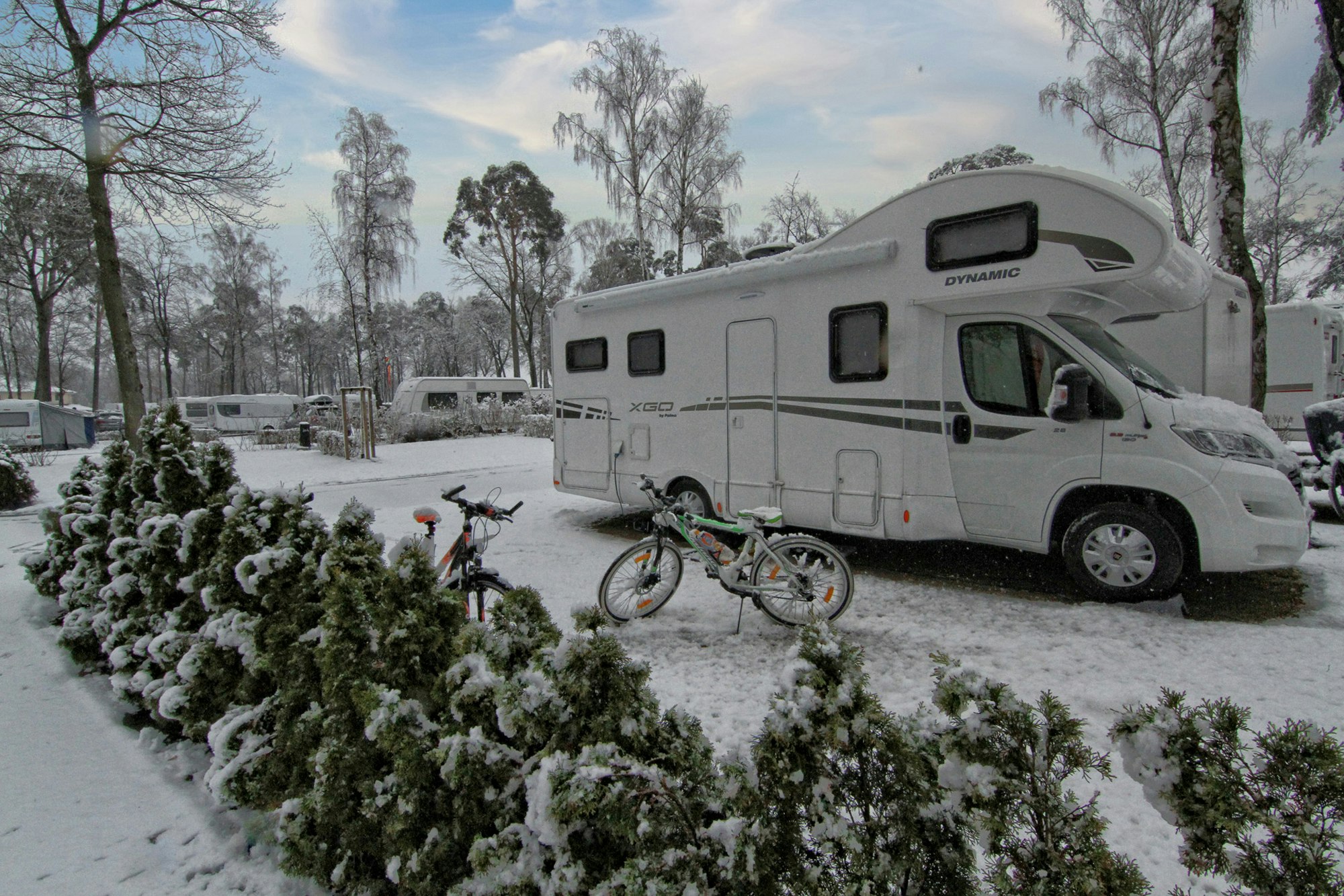 Weißes Wohnmobil auf einem Stellplatz im Schnee, davor Fahrräder, Hecken und Bäume am Campingplatz.