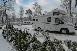 Weißes Wohnmobil auf einem Stellplatz im Schnee, davor Fahrräder, Hecken und Bäume am Campingplatz.