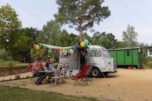 Wohnmobil mit Vorzelt und bunten Wimpeln auf dem Campingplatz, daneben Tisch und Stühle unter Bäumen.
