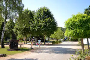 Weg im Campinggelände mit Bäumen, Spielgeräten und zwei Personen, im Hintergrund ein Wohnwagen und ein Gebäude.