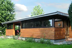Holzbungalow mit großer Terrasse, Holzgeländer und Fenstern, umgeben von Bäumen und Blumen im Garten.