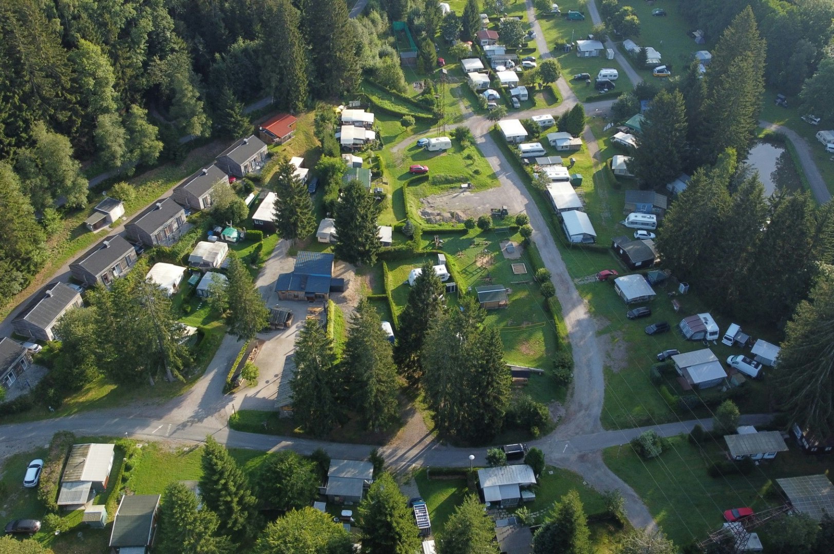 Luftaufnahme einer Campinganlage mit Wohnwagen, Stellplätzen, Wegen und mehreren Gebäuden inmitten von Bäumen und Grünflächen.