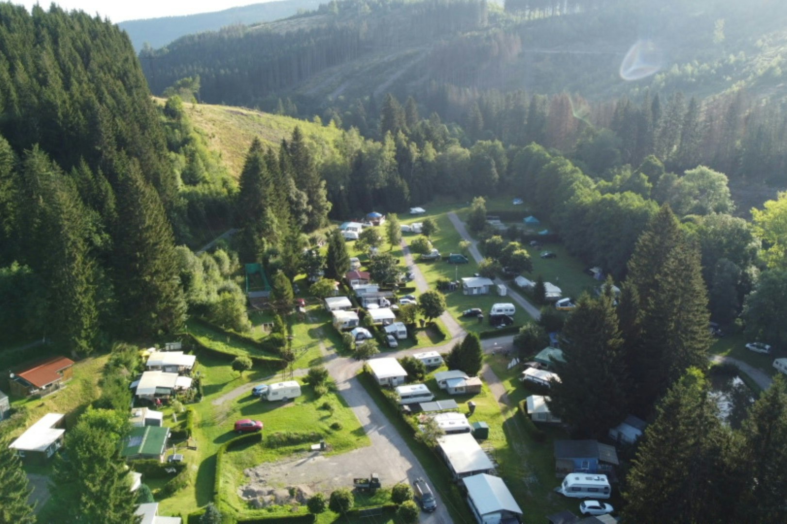 Campingplatz im grünen Tal mit vielen Stellplätzen, Wohnmobilen und Autos, umgeben von dichten Nadelwäldern und Bergen.