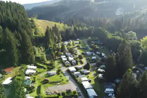Campingplatz im grünen Tal mit vielen Stellplätzen, Wohnmobilen und Autos, umgeben von dichten Nadelwäldern und Bergen.