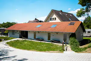 Campinggebäude mit rotem Ziegeldach, gepflastertem Weg und Rasenfläche vor dem Haus, daneben ein Wohnwagen.