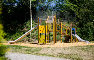 Spielplatz mit Klettergerüst und Rutsche auf einem Campingplatz, umgeben von Bäumen und Grasfläche.