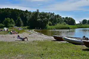 Ruderboot am Ufer eines Flusses, daneben Gras und Steinschüttung; im Hintergrund sitzen Menschen am Strand.