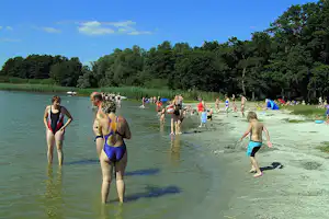 Badegäste im See am Campingplatz, flacher Uferbereich mit Sandstrand und Bäumen im Hintergrund.