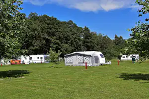 Wohnwagen und Zelt auf einem grünen Campingplatz, umgeben von Bäumen, mit Rasenfläche und Stellplätzen.