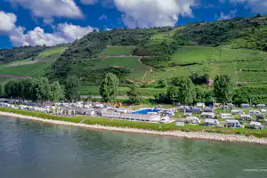 Campingplatz am Fluss mit Stellplätzen, Wohnmobilen und einem Gebäude am Ufer, umgeben von Weinbergen und Hügeln.