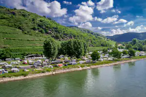Campingplatz am See mit vielen Wohnwagen und Zelten, Rasenflächen, Bäumen und Blick auf grüne Hügel und Berge.