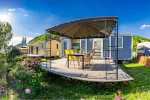 Mobilheim auf Holzterrasse mit Markise, Tisch und gelben Stühlen, umgeben von Blumenbeeten und Rasen, im Hintergrund weitere Unterkünfte.