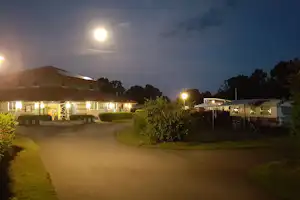 Campingplatz bei Dämmerung: beleuchteter Weg zwischen Wohnwagen und Bungalows, Bäume und Hecken am Rand.