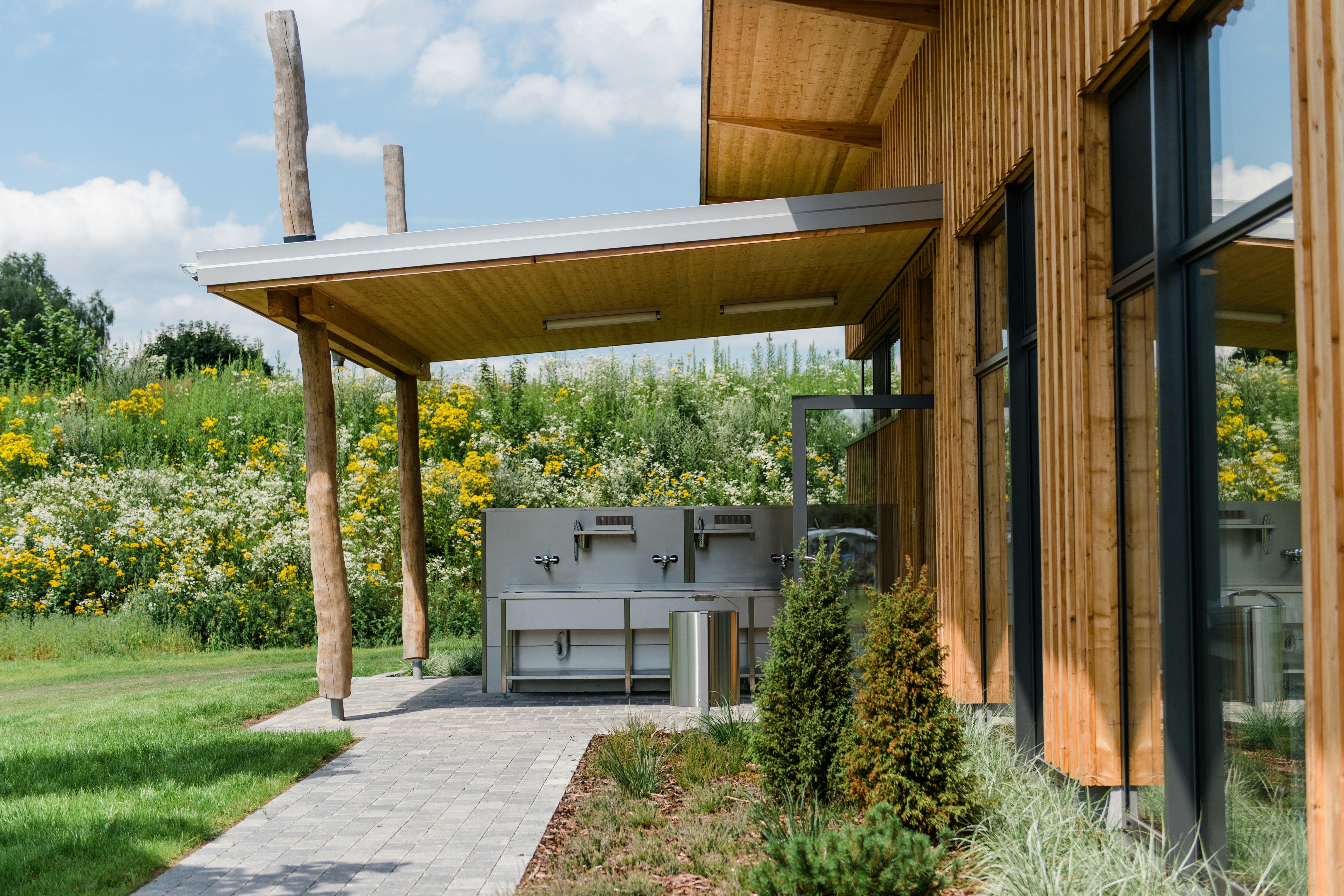 Holzhaus mit Grillbereich und überdachtem Unterstand, gepflasterter Weg, Rasen und Blumenbeete im Garten.
