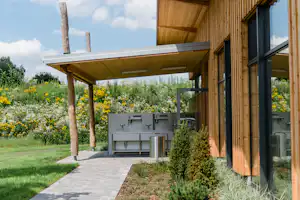 Holzhaus mit Grillbereich und überdachtem Unterstand, gepflasterter Weg, Rasen und Blumenbeete im Garten.