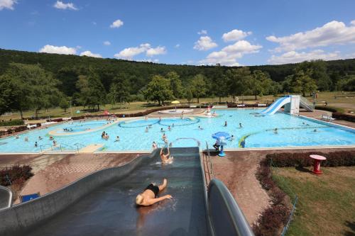 Großes Freibad mit Rutschen und vielen Badegästen, umgeben von Rasenflächen und Bäumen vor bewaldeten Hügeln.