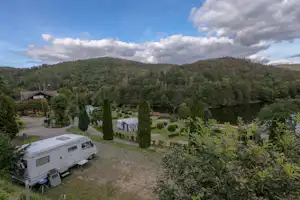 Wohnwagen auf einem Campingplatz mit gepflegten Hecken, Gartenwegen und mehreren Fahrzeugen, im Hintergrund bewaldete Hügel und Berge.