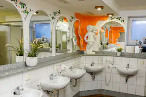 Camping-Badezimmer mit mehreren Waschbecken, Spiegeln und weißen Fliesen, orangefarbener Wand und dekorativen Bögen mit Pflanzen.