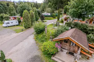 Holzhütte mit Terrasse, gepflegter Gartenanlage und Wohnwagen auf dem Campingplatz, umgeben von Bäumen und Wegen.