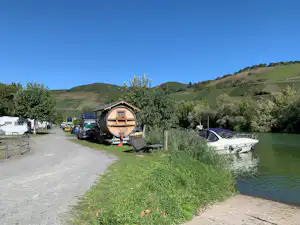 Holzhaus am Seeufer mit Wohnwagen, Auto und Kiesweg; im Hintergrund Wiesen und Hügel unter klarem Himmel.