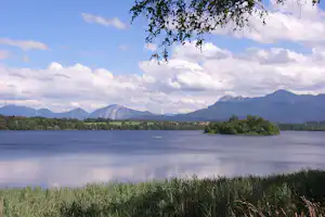 See mit ruhiger Wasseroberfläche, im Vordergrund Gras, dahinter Uferwald und Berge unter wolkigem Himmel.