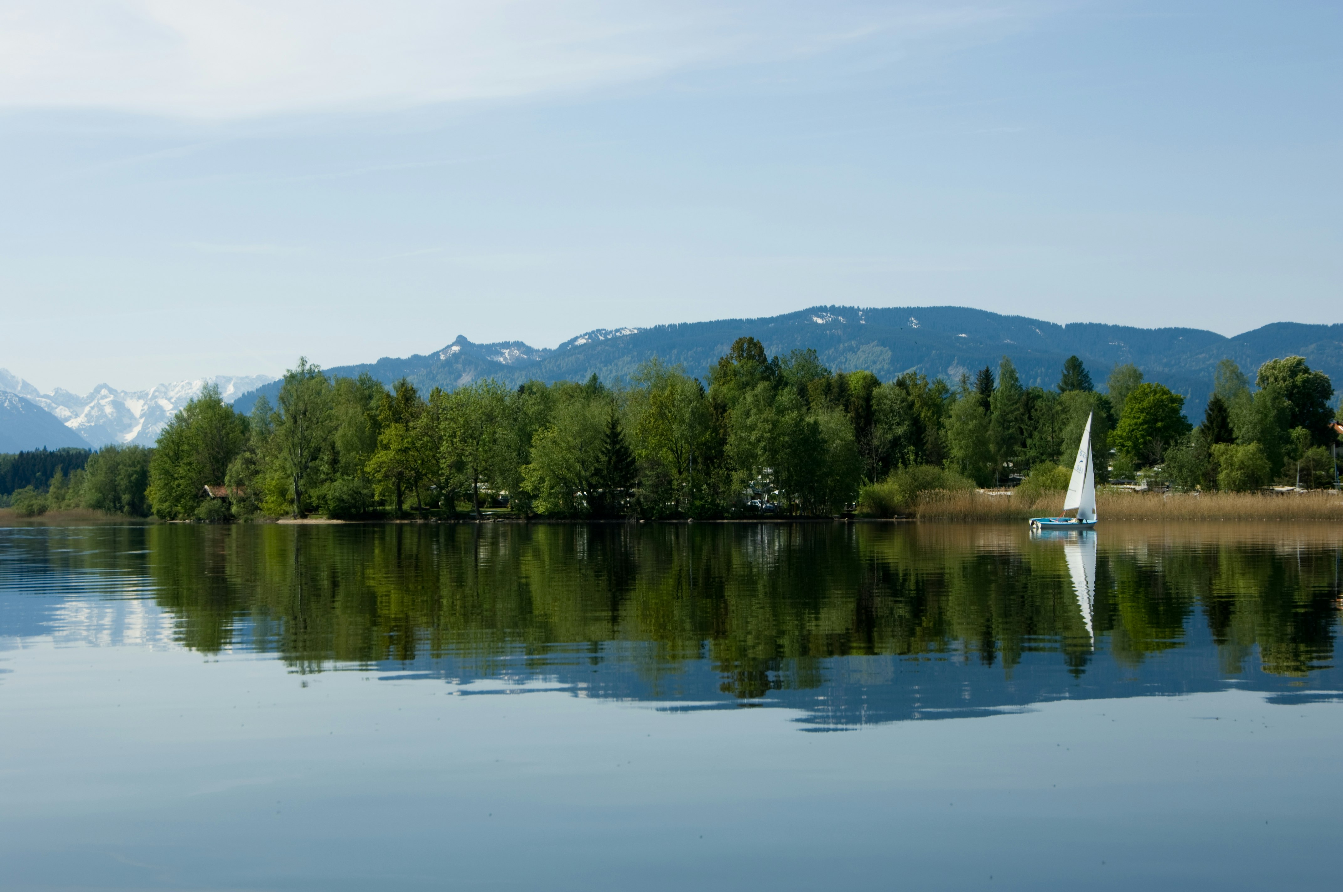 See mit Segelboot am Ufer, Bäume im Vordergrund und Berge in der Ferne, Spiegelung im Wasser.