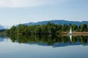 See mit Segelboot am Ufer, Bäume im Vordergrund und Berge in der Ferne, Spiegelung im Wasser.