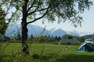 Wohnwagen auf Campingwiese, umgeben von Bäumen und Zaun, im Hintergrund Berge und ein Zelt.