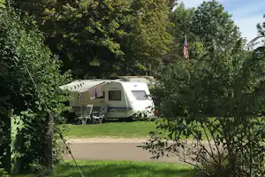 Wohnwagen auf einem Campingplatz im Grünen, mit Markise, Tisch und Stühlen vor dichtem Buschwerk und Bäumen.