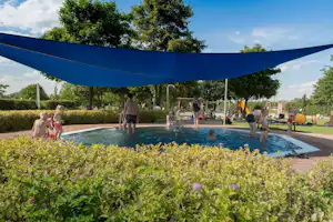 Camping-Pool mit Sonnensegel, Kinder und Erwachsene im Wasser, Liegen und Wege im Hintergrund, Bäume und Hecken.