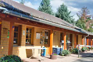 Holzbungalow am Campingplatz mit Terrasse, Sitzgelegenheiten und Blumenkübeln vor den Eingängen, unter einem großen Dachvorsprung.