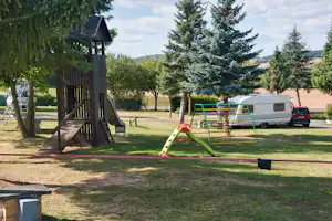 Spielplatz mit Rutsche und Holzspielturm auf dem Campingplatz, im Hintergrund Wohnwagen und Auto, Bäume und Rasen.