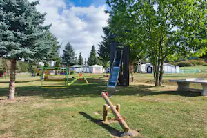 Spielplatz auf dem Campingplatz mit Klettergerüst, Rasenfläche und Bäumen, im Hintergrund Zelte und Wohnwagen.
