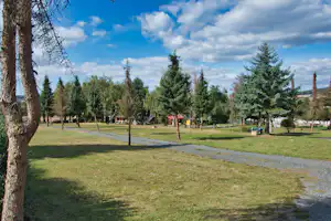 Grüne Campinganlage mit Rasenflächen, Wegen und vielen Bäumen, im Hintergrund weitere Stellplätze und kleine Gebäude.