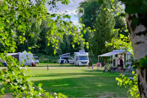 Campingplatz im Grünen: Wohnwagen und Zelte auf Stellplätzen, Tische und Stühle unter Sonnenschirmen, umgeben von Bäumen.