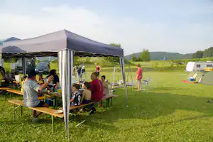 Essenszelt auf dem Campingplatz mit Holztischen, Familiengruppe am Tisch, Autos und Wohnwagen im Hintergrund, Wiese und Bäume.