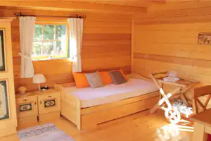 Gemütliches Holzschlafzimmer im Campinghaus: Doppelbett mit Kissen, Nachttisch, Tisch und Stuhl, Fenster mit Vorhängen.